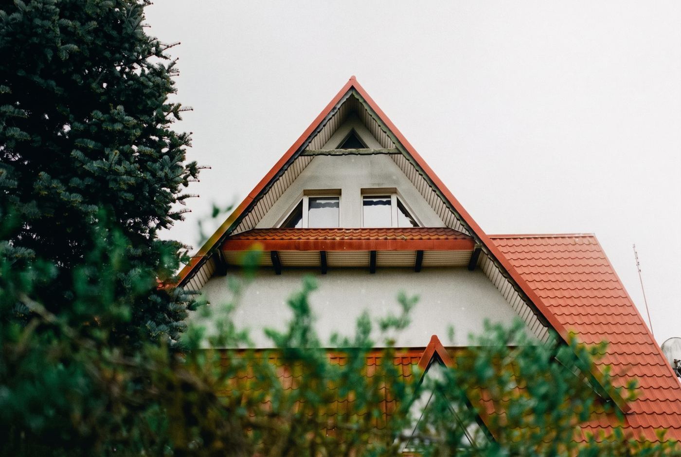 The gable and red tiled roof of a typical Polish suburban house, framed by conifers on an overcast day.