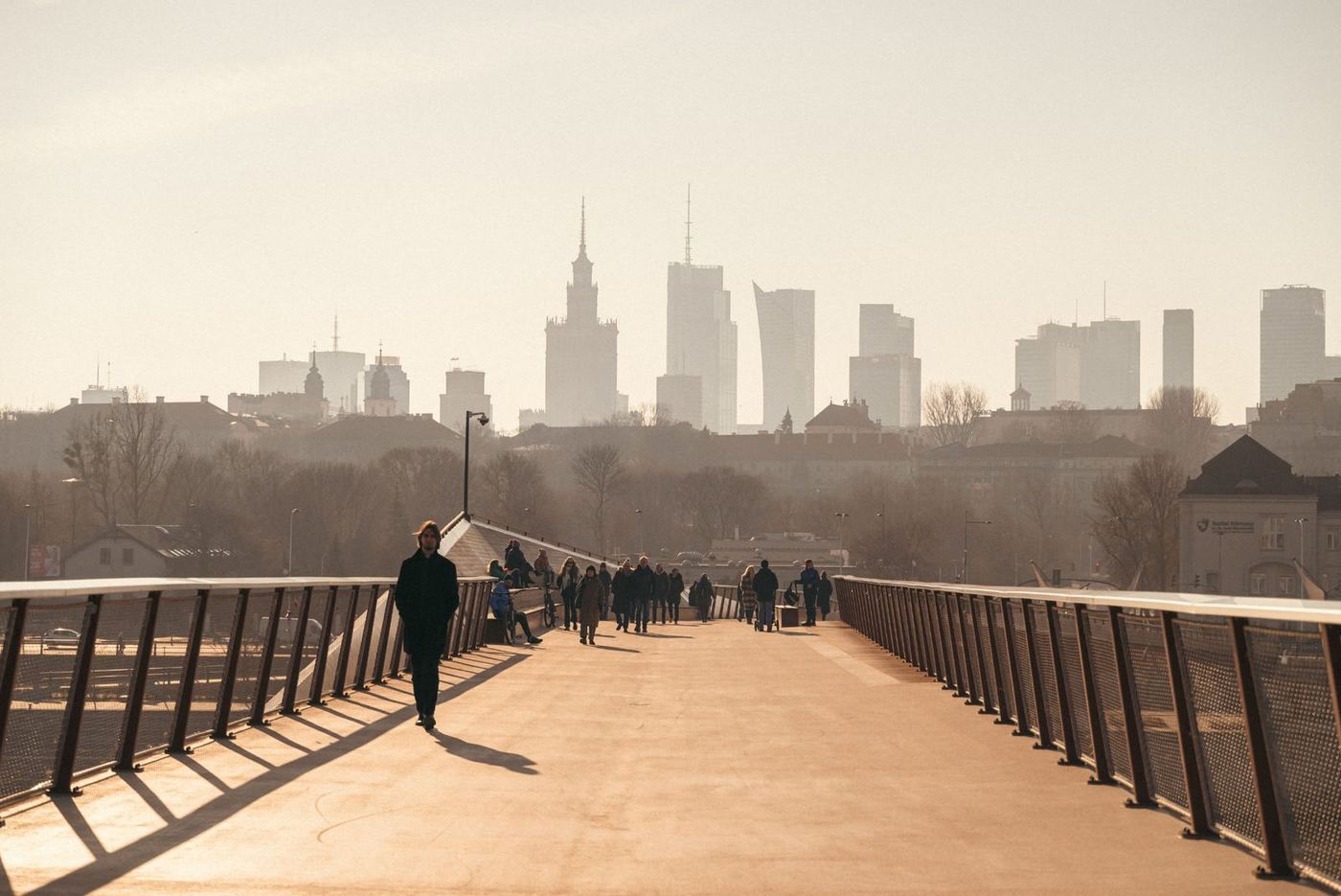 A warm-toned view of a pedestrian bridge in Warsaw, with people walking along the wide deck and the Palace of Culture and Science rising above the hazy city skyline in the background.