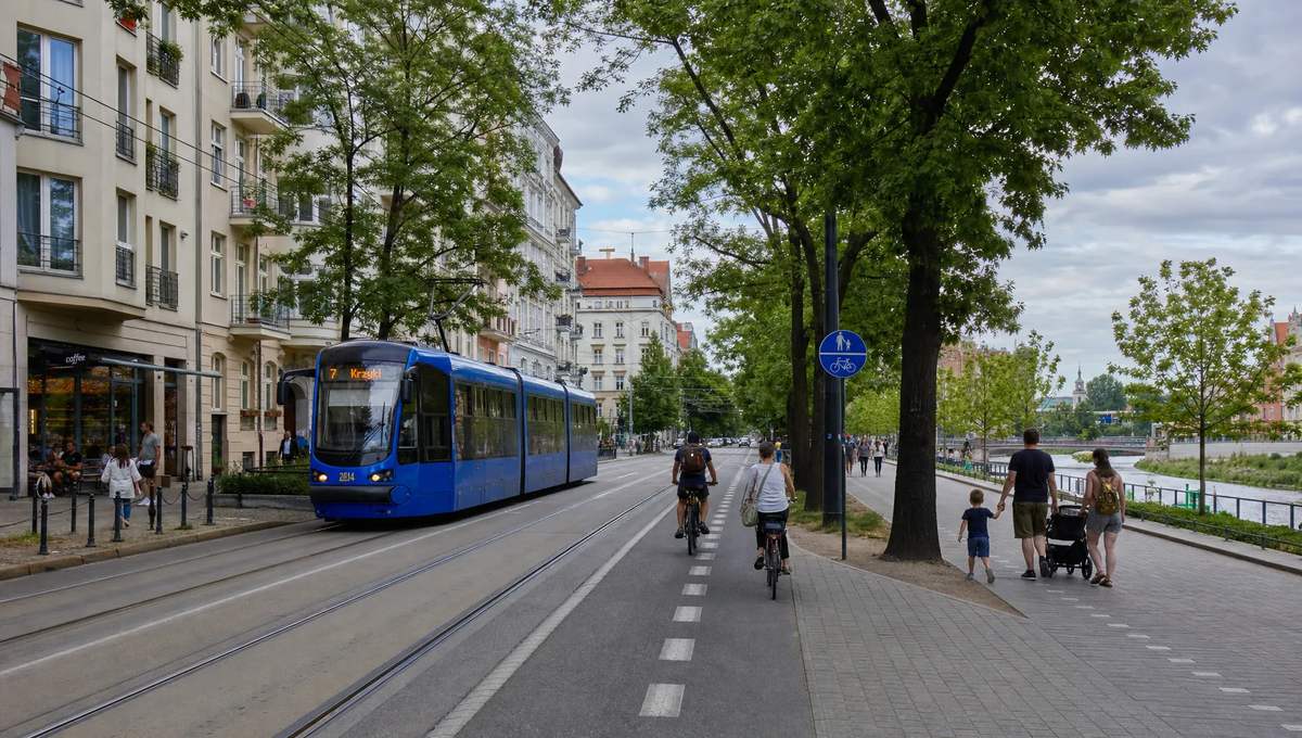 A calm residential street in Wrocław with tram tracks, leafy trees, apartment buildings, cyclists, and families walking near a riverside park, showing everyday life rather than tourism.